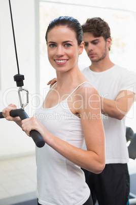 Fit smiling woman using weights machine for arms with her traine