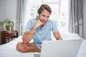 Handsome man sitting on bed using laptop smiling at camera
