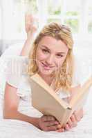 Pretty blonde lying on bed smiling at camera with book