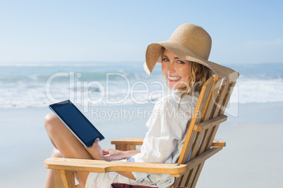 Smiling blonde sitting on wooden deck chair by the sea using tab