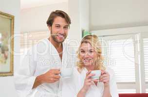 Cute couple in bathrobes smiling at camera together holding cups