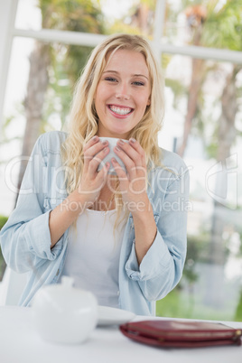 Pretty blonde sitting at table having coffee smiling at camera