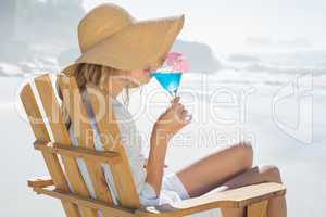 Smiling blonde relaxing in deck chair by the sea sipping cocktai