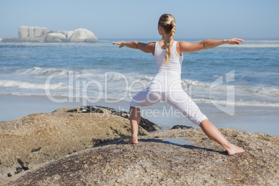 Blonde woman standing in warrior pose on beach on rock