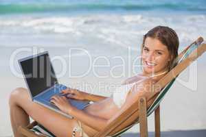 Smiling woman relaxing in deck chair on the beach using laptop