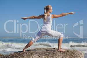 Blonde woman standing in warrior pose on beach on rock