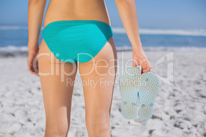 Rear view of fit woman in bikini on beach holding flip flops