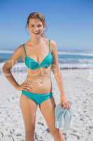 Fit smiling woman in bikini on beach holding flip flops