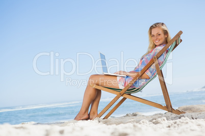 Blonde sitting on beach using her laptop smiling at camera