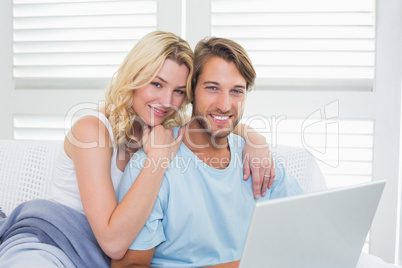 Happy casual couple sitting on couch using laptop smiling at cam