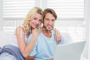 Happy casual couple sitting on couch using laptop smiling at cam