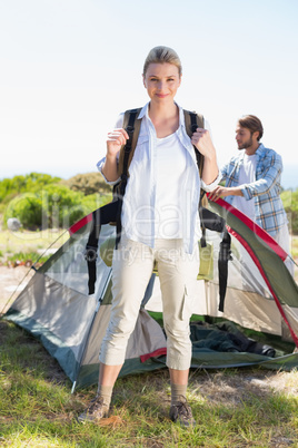 Attractive blonde smiling at camera while partner pitches tent