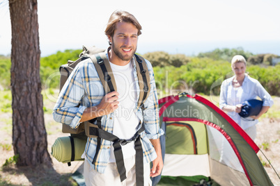 Attractive man smiling at camera while partner pitches tent
