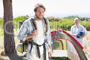 Attractive man smiling at camera while partner pitches tent