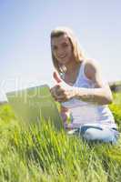 Pretty blonde sitting on grass using her laptop smiling at camer