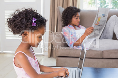 Cute daughter using laptop at desk with mother on couch