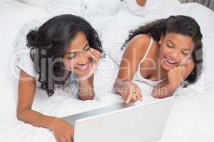 Smiling mother and daughter using laptop together on bed