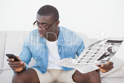 Casual man sitting on sofa texting on phone holding the paper