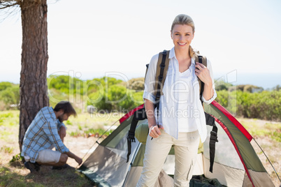 Attractive blonde smiling at camera while partner pitches tent