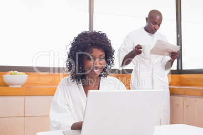 Pretty woman in bathrobe using laptop at table with partner in b