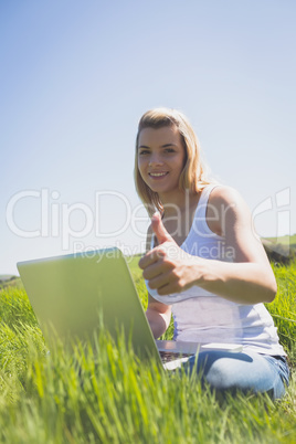 Pretty blonde sitting on grass using her laptop smiling at camer