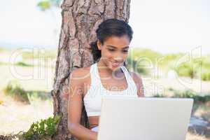 Fit woman sitting against tree using laptop