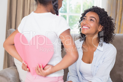 Pretty mother sitting on couch with daughter hiding heart card