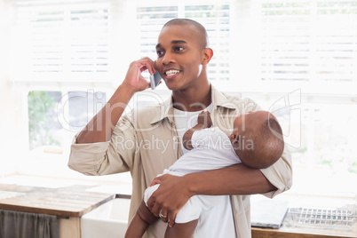 Happy father feeding his baby boy a bottle while on the phone