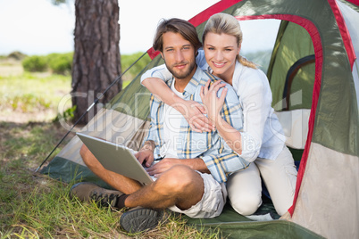 Attractive couple sitting by their tent using laptop smiling at