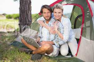 Attractive couple sitting by their tent using laptop smiling at