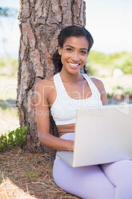 Fit woman sitting against tree using laptop