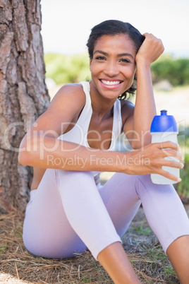 Fit woman sitting against tree holding water bottle