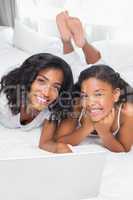 Smiling mother and daughter using laptop together on bed
