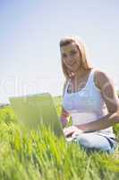 Pretty blonde sitting on grass using her laptop smiling at camer