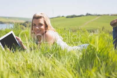 Pretty blonde lying on grass using her tablet smiling at camera