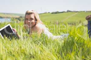 Pretty blonde lying on grass using her tablet smiling at camera