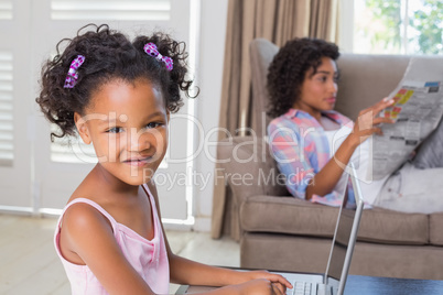 Cute daughter using laptop at desk with mother on couch