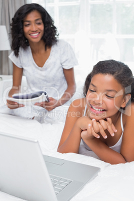 Mother reading magazine with daughter using laptop on bed