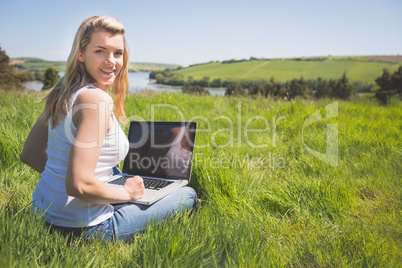 Pretty blonde sitting on grass using her laptop smiling at camer