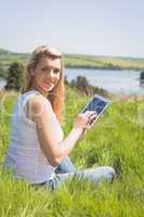 Pretty blonde sitting on grass using her tablet smiling at camer