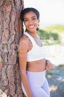 Fit woman leaning against tree smiling at camera