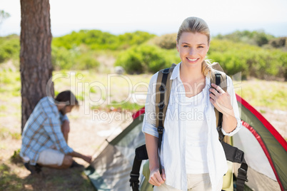 Attractive blonde smiling at camera while partner pitches tent