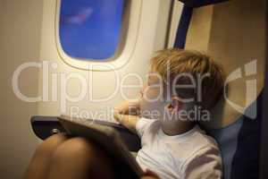 Boy in plane looking out illuminator with pad on lap