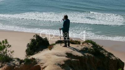 Man Stands on a Cliff Above the Ocean and Drinking Water from Bottle