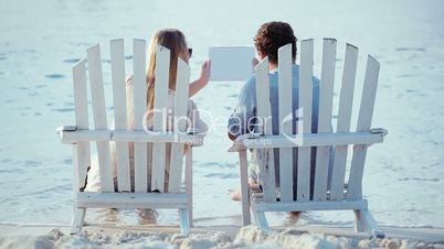 Young couple in chaise-longues holding pad and looking at it