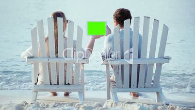Woman and man sitting on the beach looking at pad with green screen
