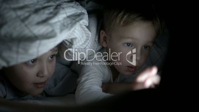 Two boys lying in bed at night watching movie on pad