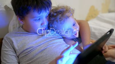 Two boys in bed with touch pad. One playing game, another watching