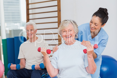 Senior woman lifting dumbbells while sitting with man and traine