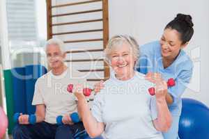 Senior woman lifting dumbbells while sitting with man and traine